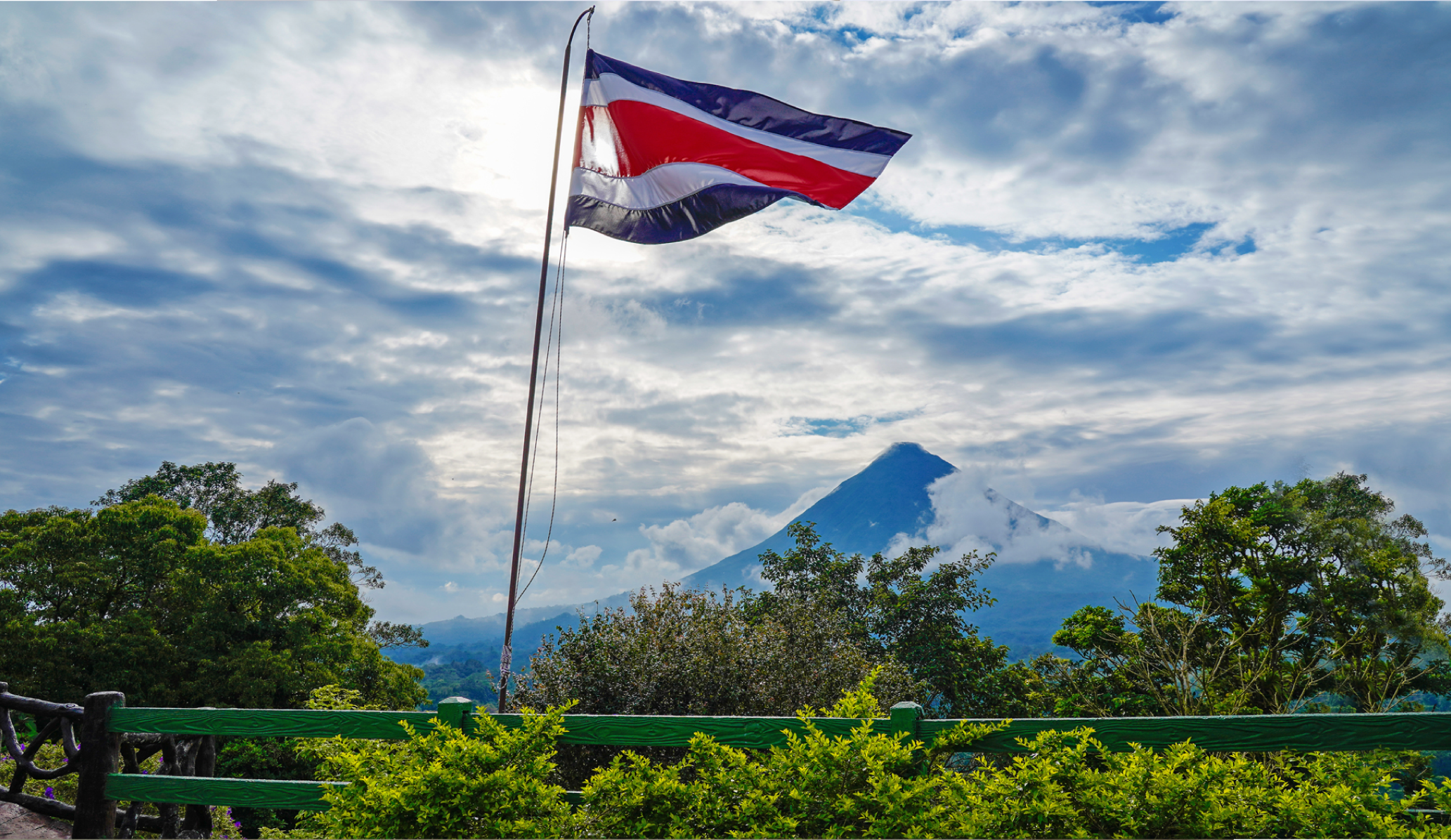 View of Costa Rica's flag waving surrounded by trees and plants, with the Arenal volcano in the background