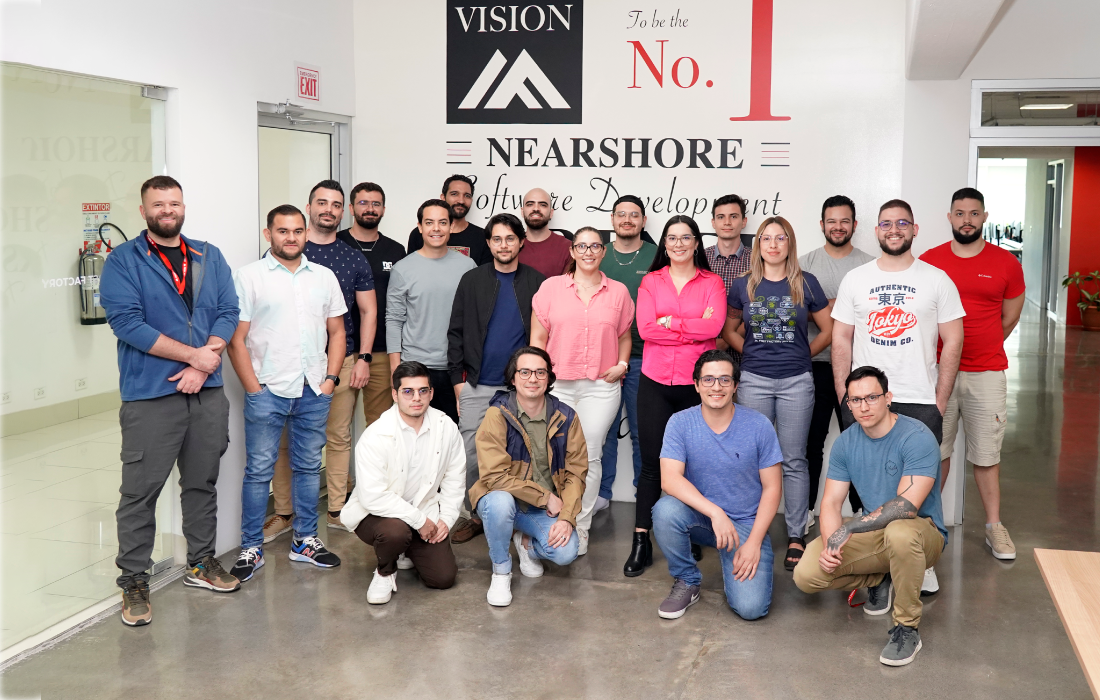 Software Engineers, QA Engineers, and Managers posing for a group photo in front of First Factory vision mural at the office