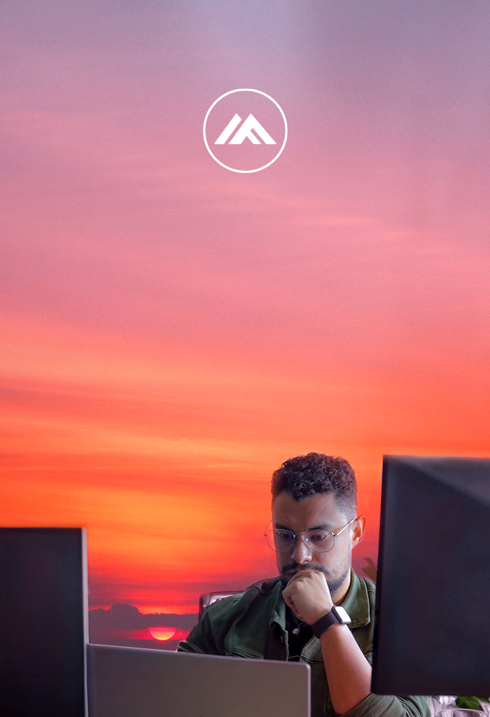 Sunset-colored background. A DevOps engineer sits, thoughtfully, in front of his computer and monitors.