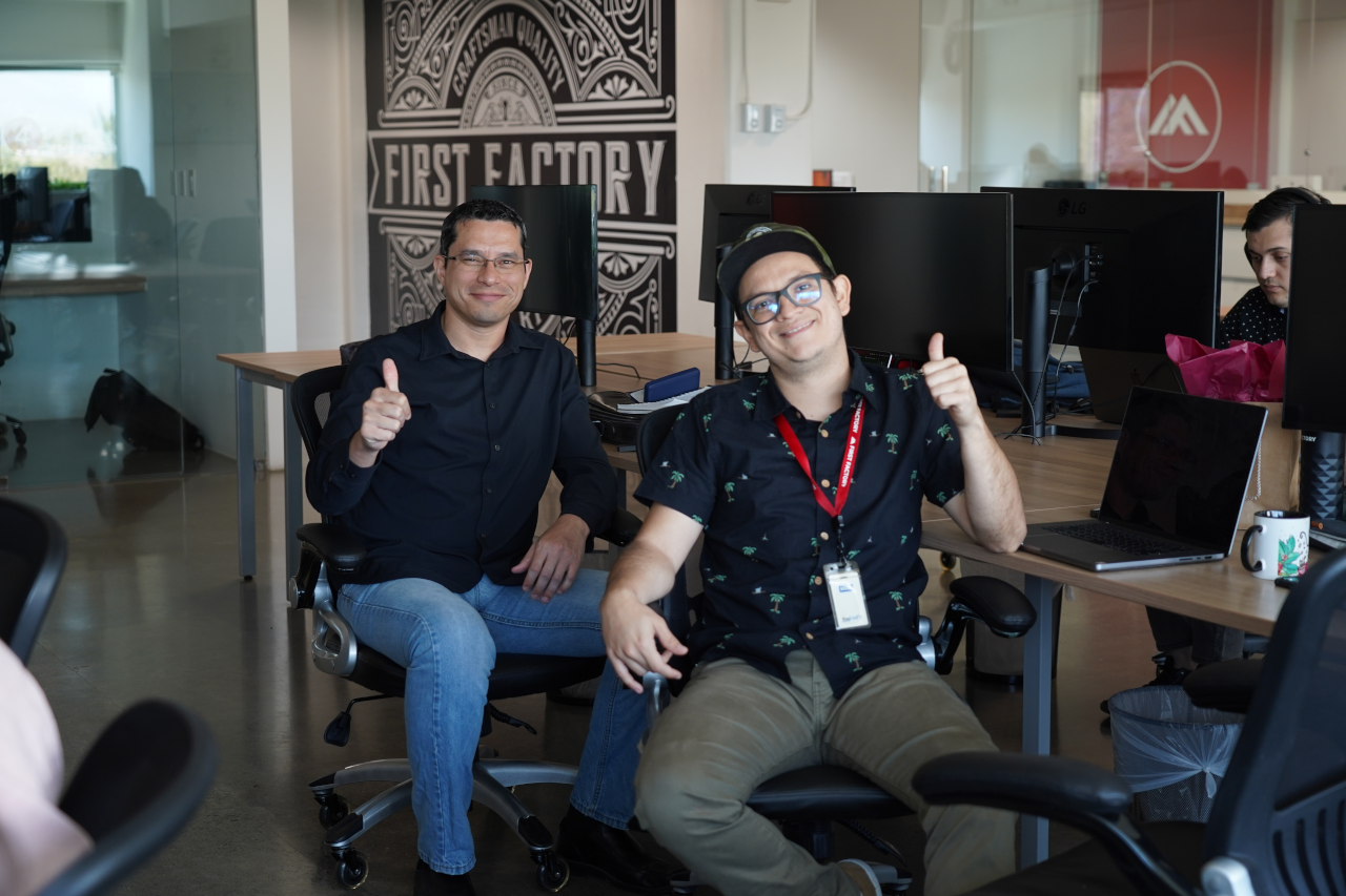 Two employees sitting at the office, smiling at the camera with thumbs up