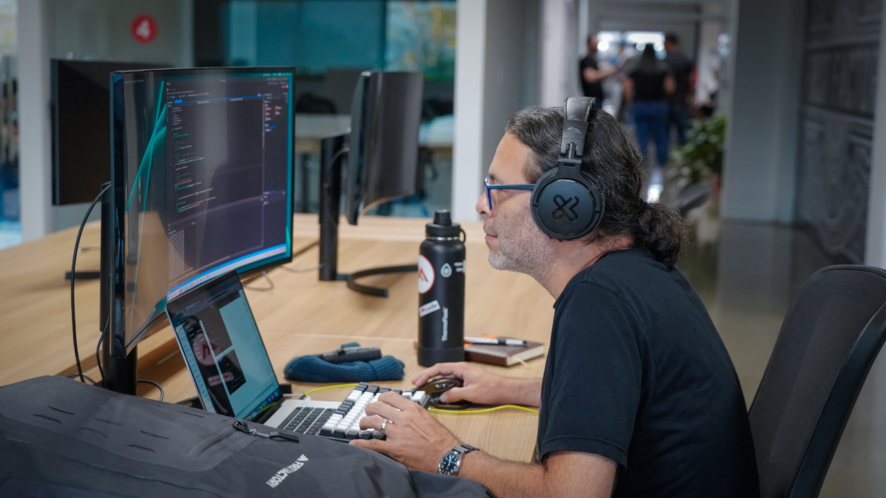 Developer wearing headphones while coding, sitting in front of the monitor with his First Factory backpack to his left